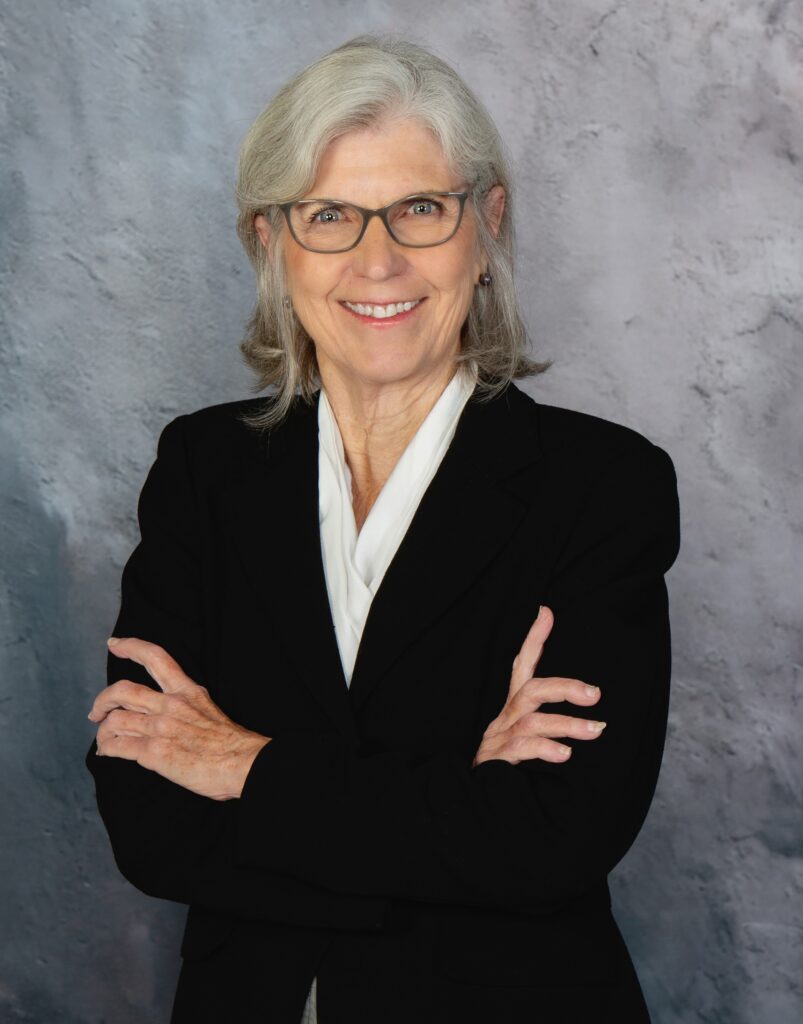 Author Marilyn F. Moriarty wearing glasses, a black blazer, and a white blouse, standing with her arms crossed in front of a gray background.